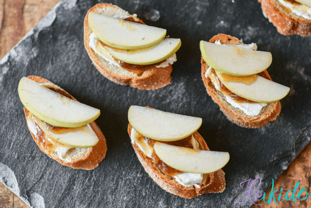 Brie, apple, and fig crostini on a slate tray with apples and figs in background