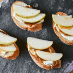 Brie, apple, and fig crostini on a slate tray with apples and figs in background