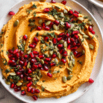 Creamy pumpkin hummus topped with pomegranate and pumpkin seeds on a white plate