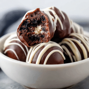 Ingredient no-bake Oreo balls in a white bowl, coated in chocolate and drizzled with white chocolate, with one ball showing the soft Oreo filling
