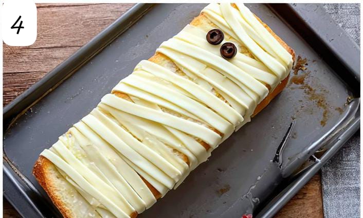 Unbaked Halloween garlic bread mummy. Cheese strips and olive eyes on bread loaf.