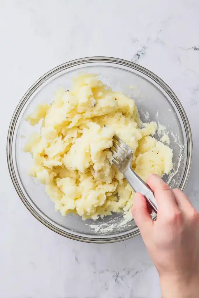 Hand mashing boiled potatoes in a clear glass bowl with a fork on a white marble background.